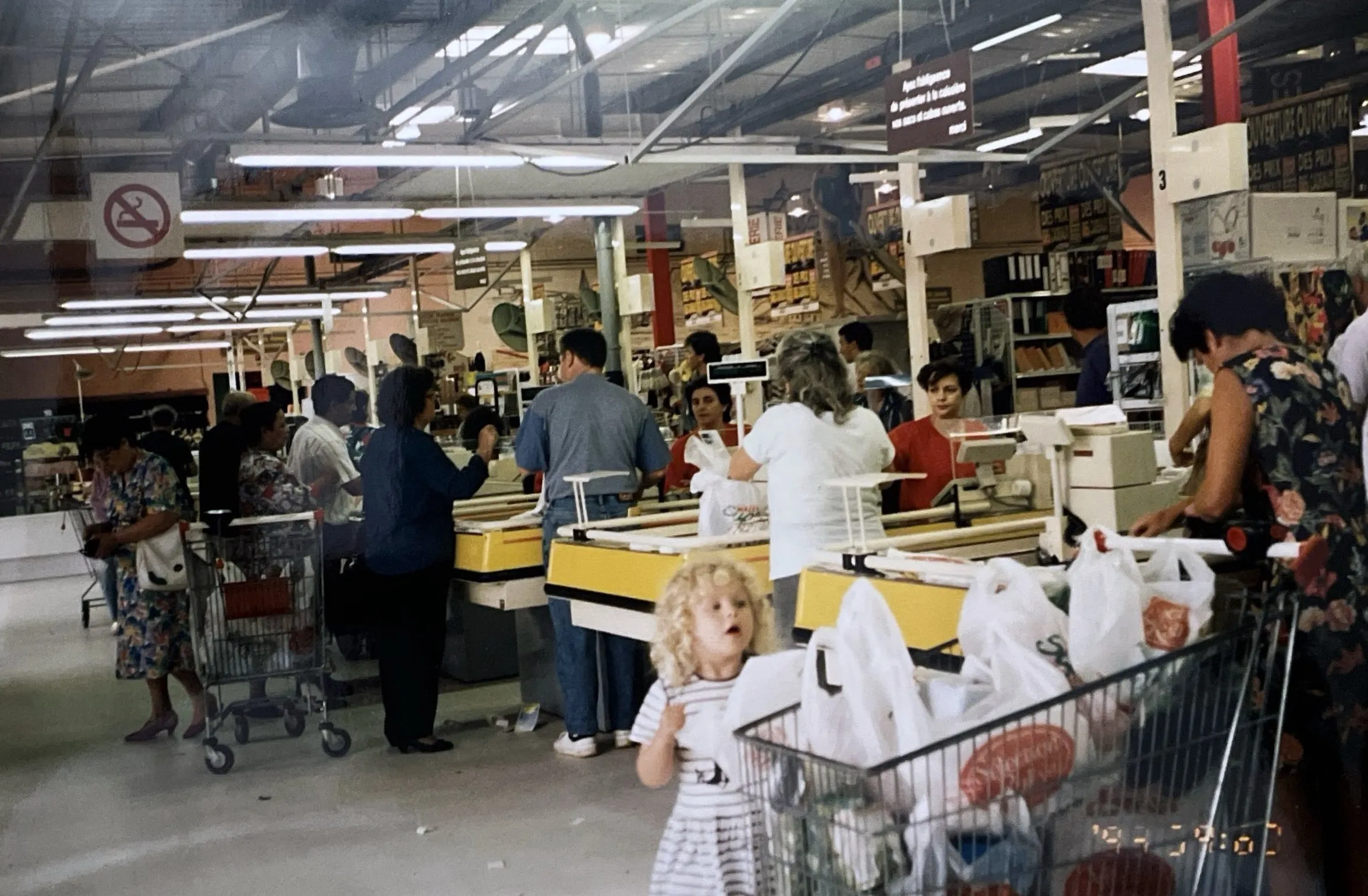 Clients aux caisses du supermarché dans les années 1990, une petite fille blonde au premier plan près d'un chariot rempli de sacs