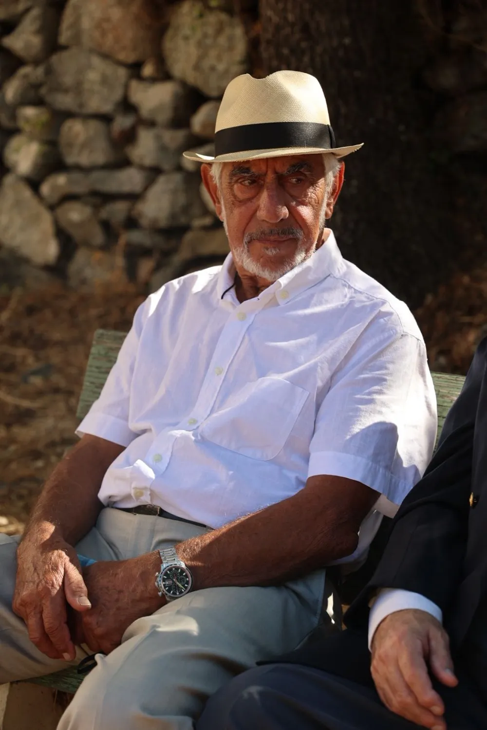 Dominique Orsini assis au soleil devant un mur de pierres sèches, chapeau de paille clair et chemise blanche