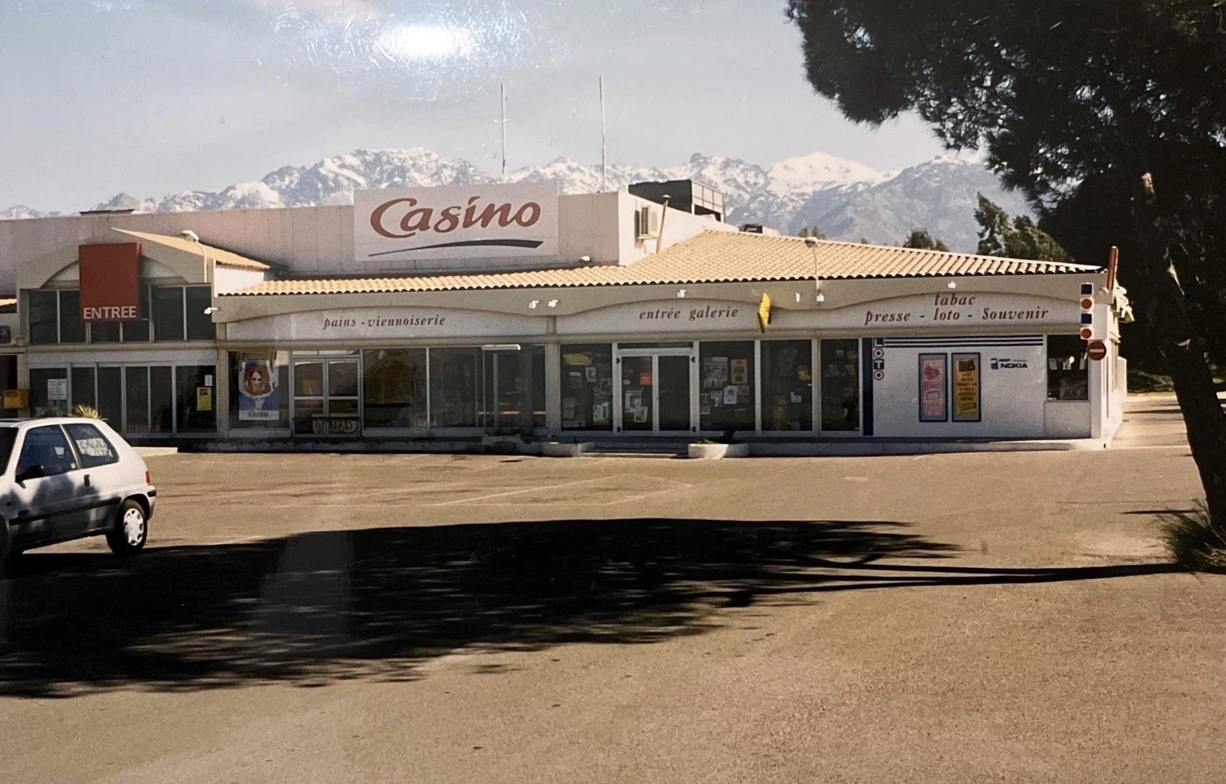 Façade du centre commercial sous l'enseigne Casino, avec l'entrée de la galerie, la boulangerie et le tabac-presse, montagnes enneigées en arrière-plan