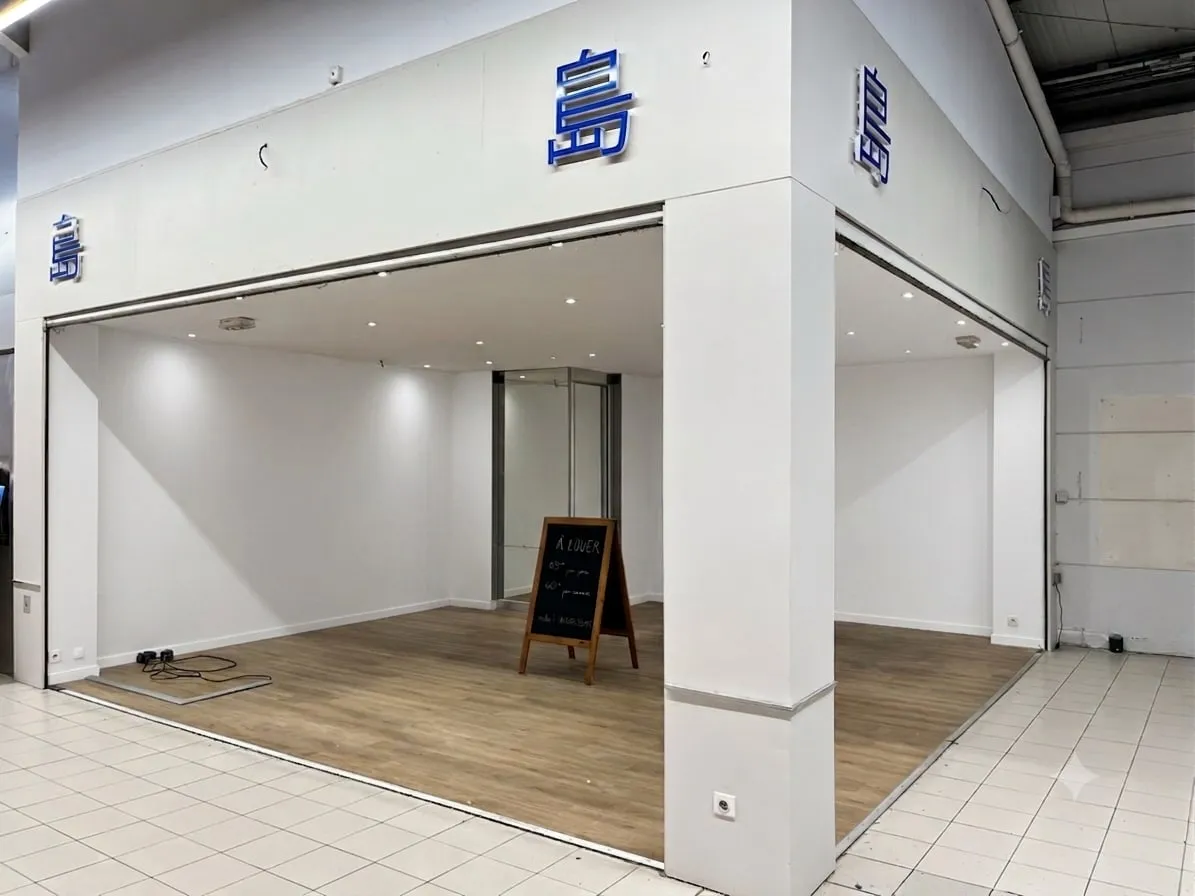 L'espace à louer dans la galerie marchande du centre commercial MAEVA : un local ouvert aux murs blancs, au sol en parquet clair, éclairé par des spots, avec une ardoise « À louer » posée au centre.