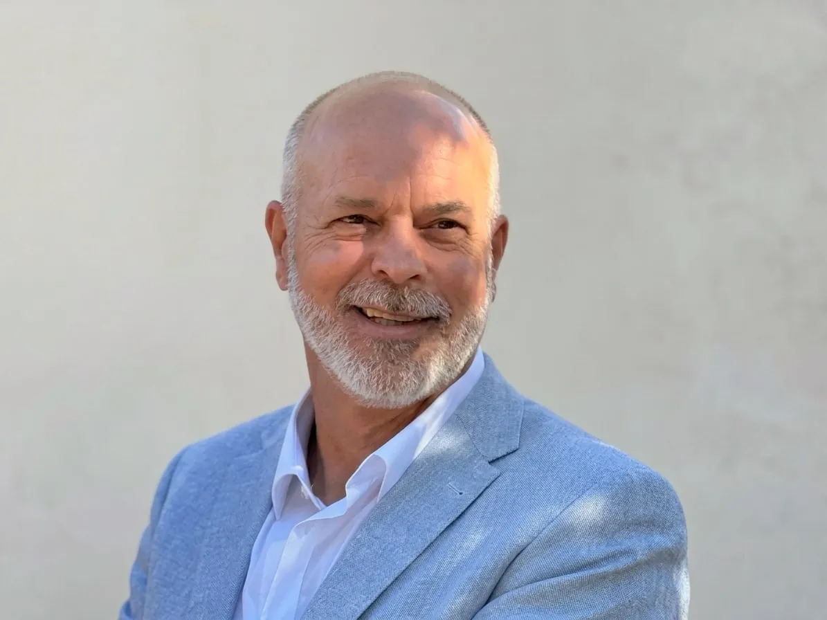 Joseph Marchetti souriant, en veste bleu clair et chemise blanche, devant un mur clair