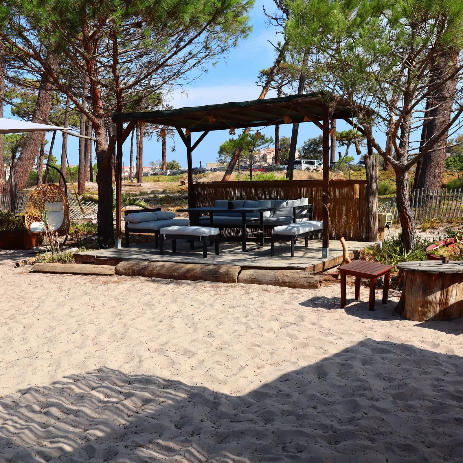 Le salon extérieur du Vol Õ Vents : canapés sous la tonnelle, parasol et pins, les pieds dans le sable